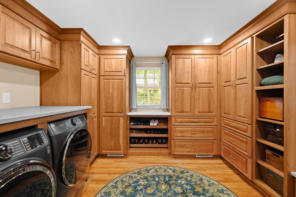 Oakmont Custom Laundry Room with Walk-In Closet Boasts Wood Cabinetry in a Warm Natural Finish, Open Shelving for Folding Items, Baskets and Decorated Touches Blending Practicality of a Laundry Room with the Elegance of a Stylish Dressing Space.